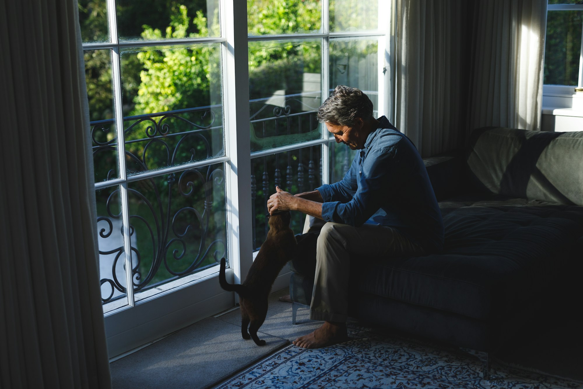 Happy senior caucasian man in living room sitting on sofa, petting his pet cat